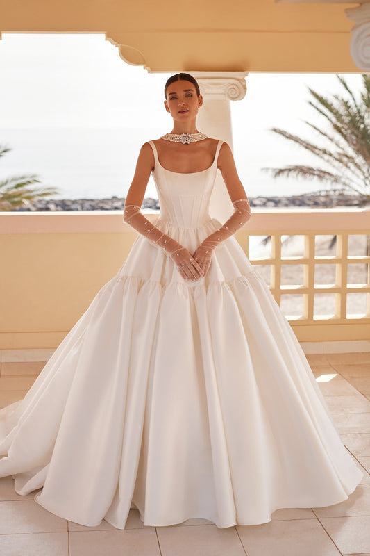 Front view of the Britania ball gown wedding dress featuring a structured square neckline, exposed boning, and a voluminous, tiered Mikado skirt with a dramatic basque waist.