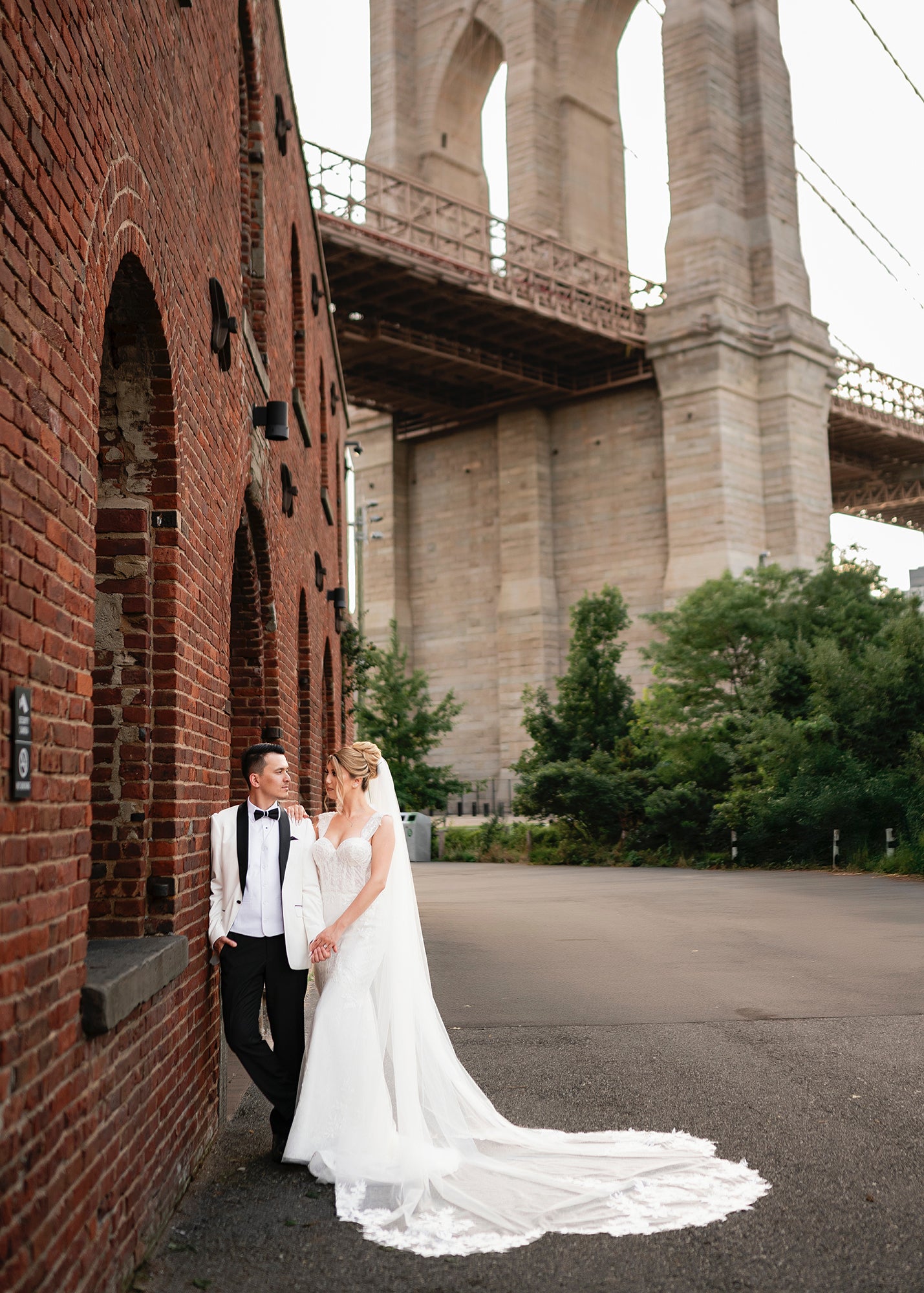 Krismil Bride Aziza in a fitted lace wedding dress with a cathedral veil, posing with her husband by the Brooklyn Bridge.