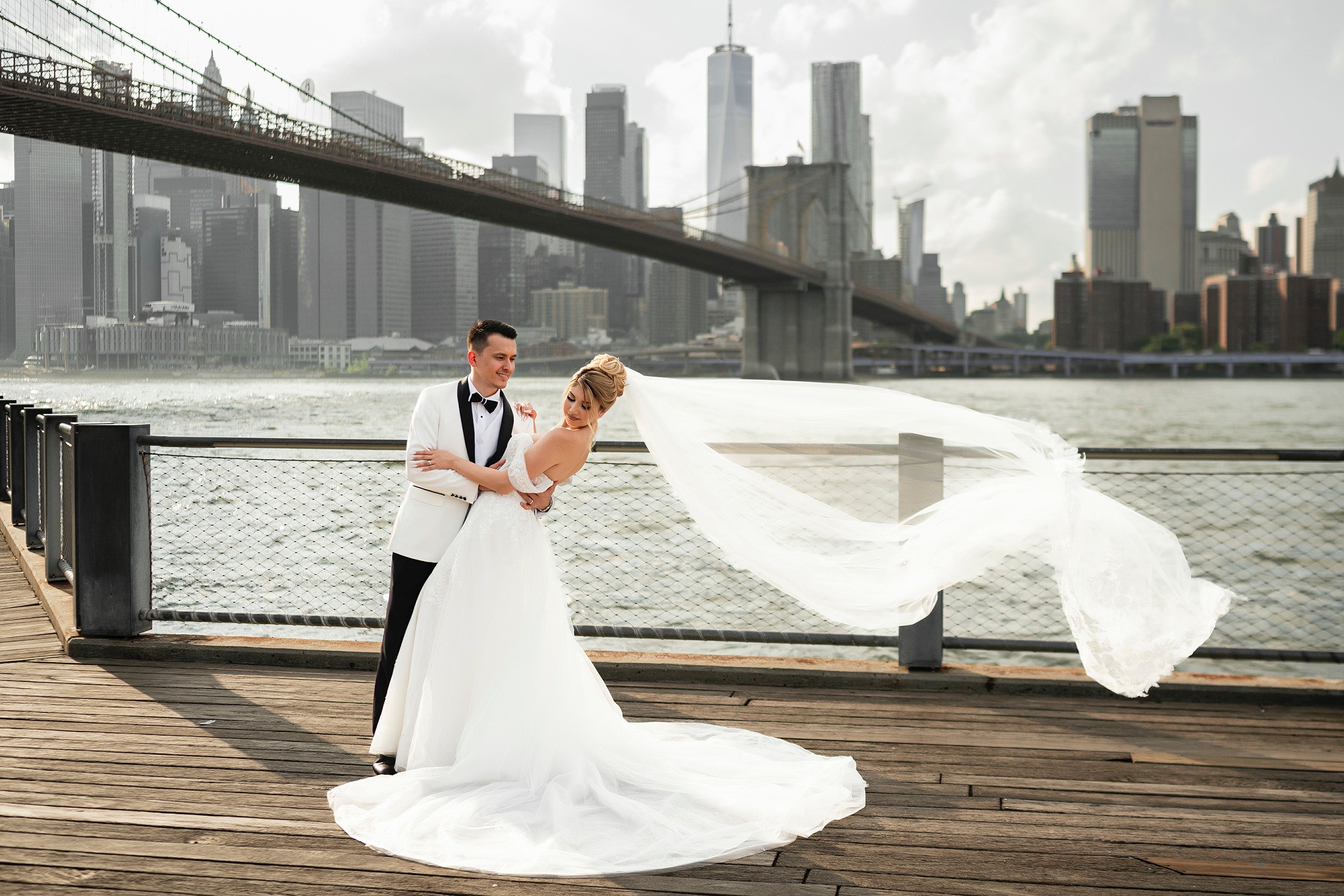 Bride and groom at Brooklyn Bridge Park, the bride wearing an off-the-shoulder A-line wedding dress with a flowing veil from Krismil Bridal, overlooking the NYC skyline
