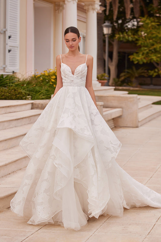 Front view of the Arran wedding dress featuring a plunging neckline, thin spaghetti straps, a draped floral jacquard bodice, and a tiered A-line skirt.