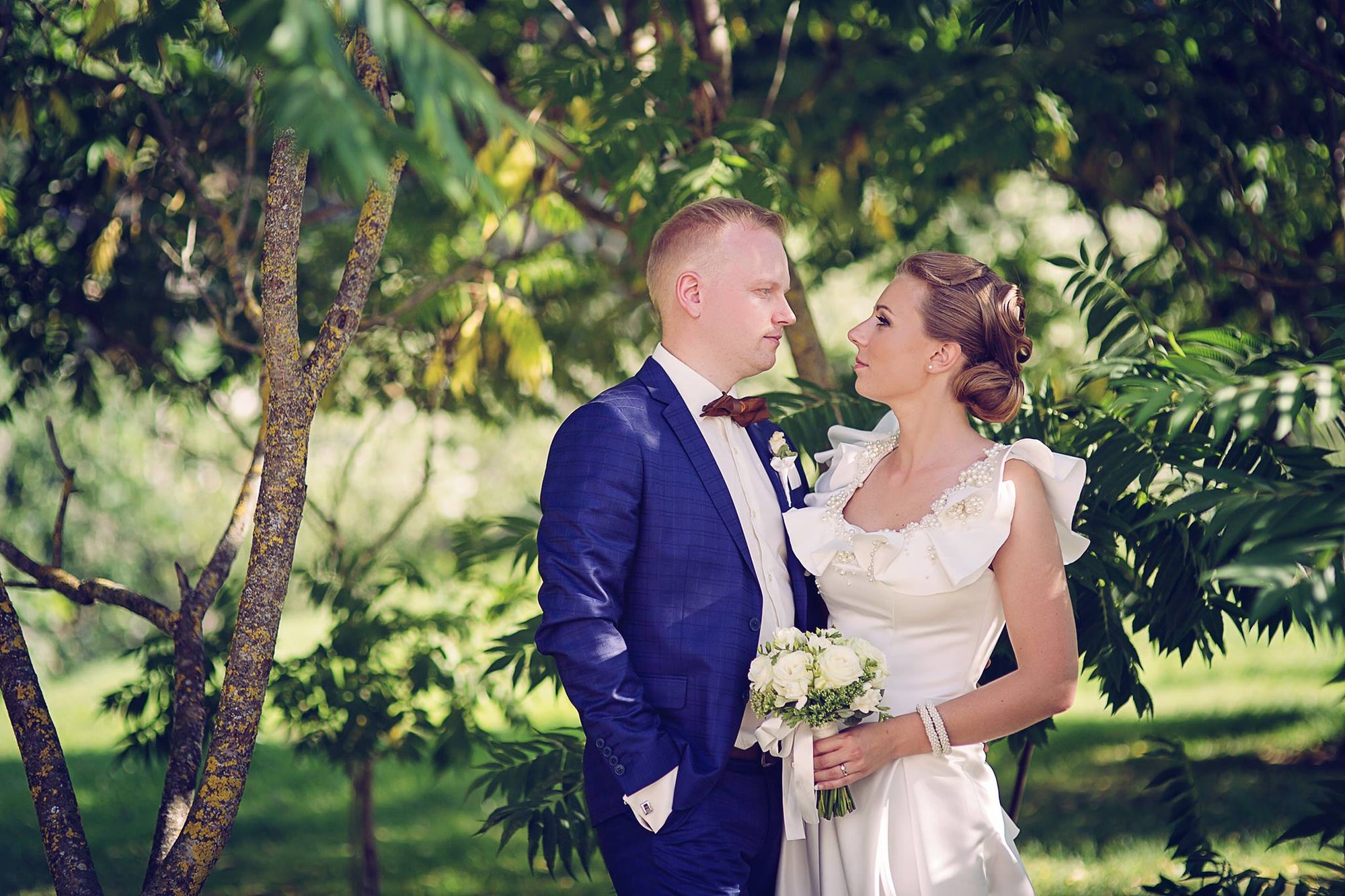 Bride in a pearl-embellished satin wedding dress by Krismil Bridal, holding a white bouquet and gazing at the groom in a blue suit during their wedding in a lush garden.