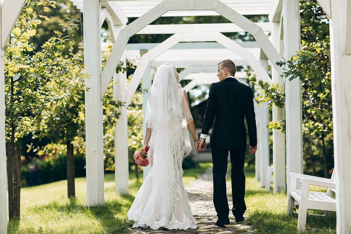 Bride in a lace mermaid wedding dress by Krismil Bridal, holding hands with the groom while walking through a garden during their wedding in Lithuania.
