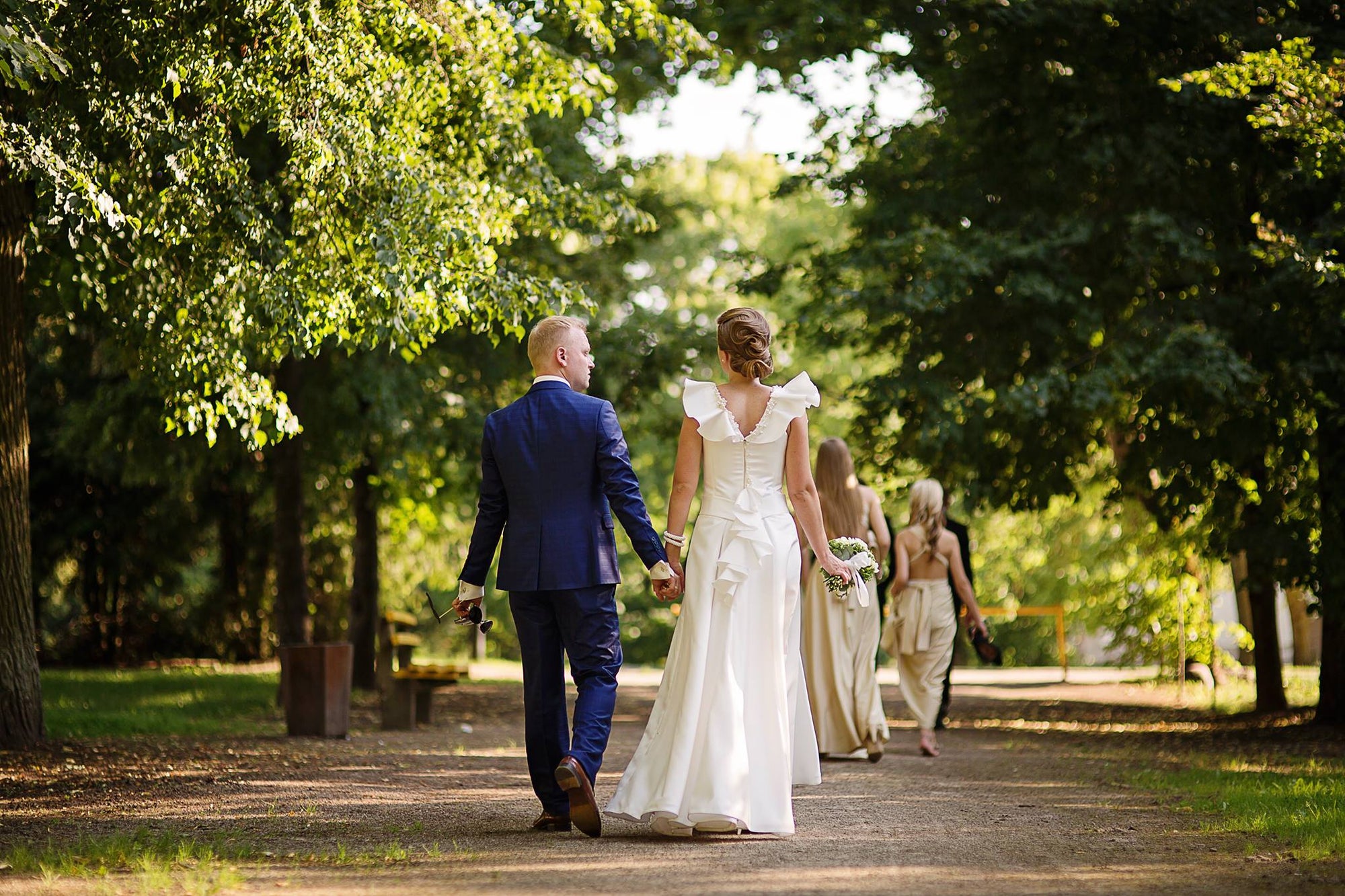 Bride in a pearl-embellished A-line wedding dress by Krismil Bridal, holding a bouquet and walking hand in hand with the groom in a blue suit through a lush garden.