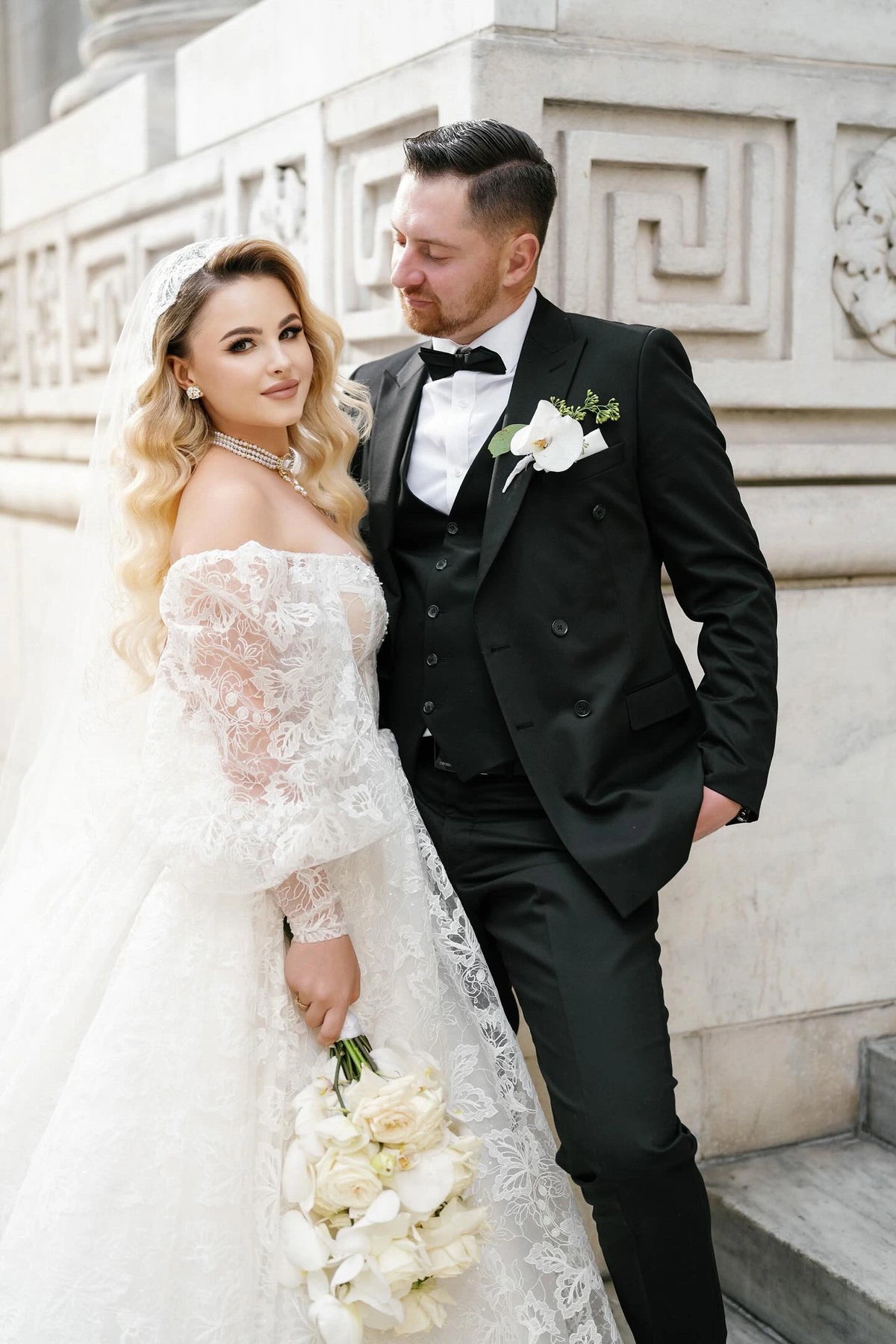 Bride Khristina in an off-the-shoulder lace wedding dress by Krismil Bridal, holding a bouquet of white roses and orchids, standing with groom Andriy in a classic black tuxedo.