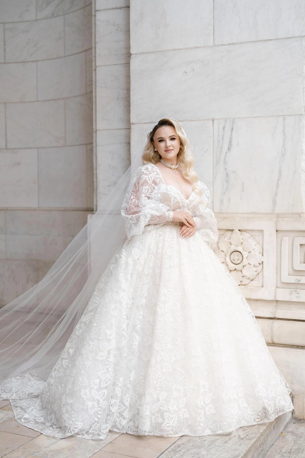 Bride Khristina in a long-sleeve princess ball gown wedding dress by Krismil Bridal, posing with a long veil and timeless beauty against a marble backdrop.