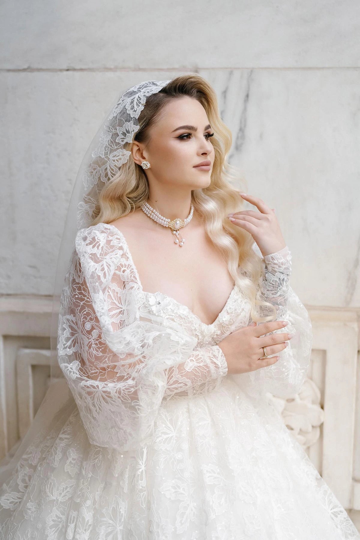 Bride Khristina in a lace wedding dress by Krismil Bridal, featuring a sweetheart neckline, sheer long sleeves, and a matching lace veil, styled with a pearl necklace and elegant waves.