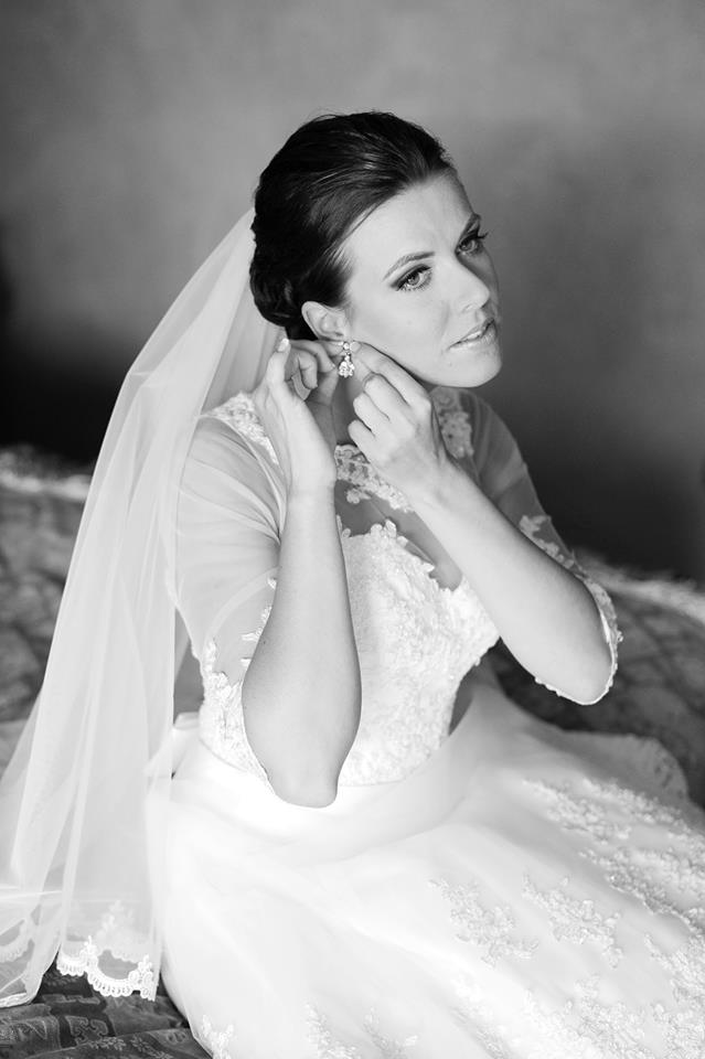Bride in a lace wedding dress by Krismil Bridal, adjusting her earrings with a serene expression, wearing a veil draped over her shoulders.