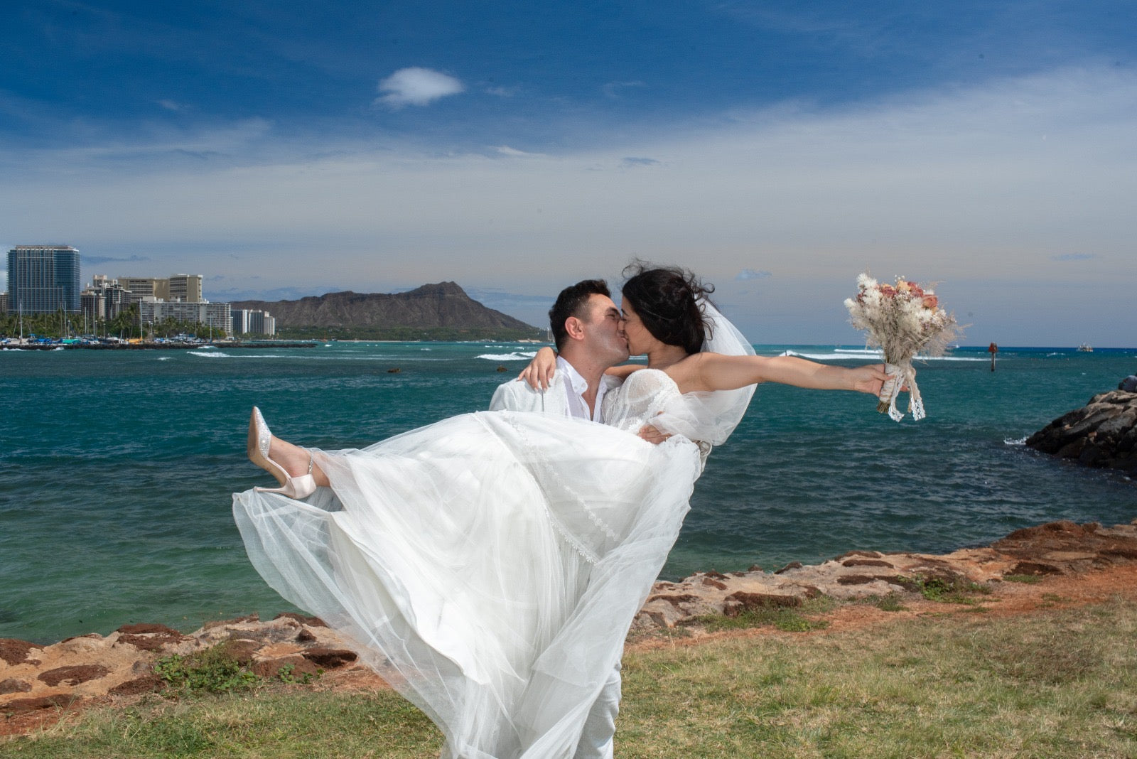 Bride Melis in a tulle wedding dress by Krismil Bridal being lifted by the groom while holding her bouquet, with the ocean and Hawaiian landscape in the background.
