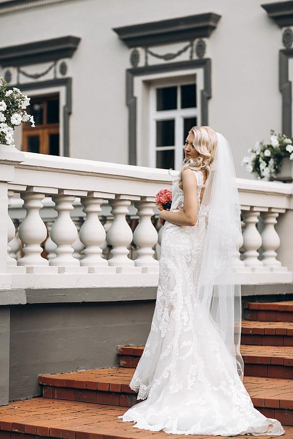 Bride in a lace mermaid wedding dress by Krismil Bridal, holding a bouquet while standing on the stairs during her wedding in Lithuania.