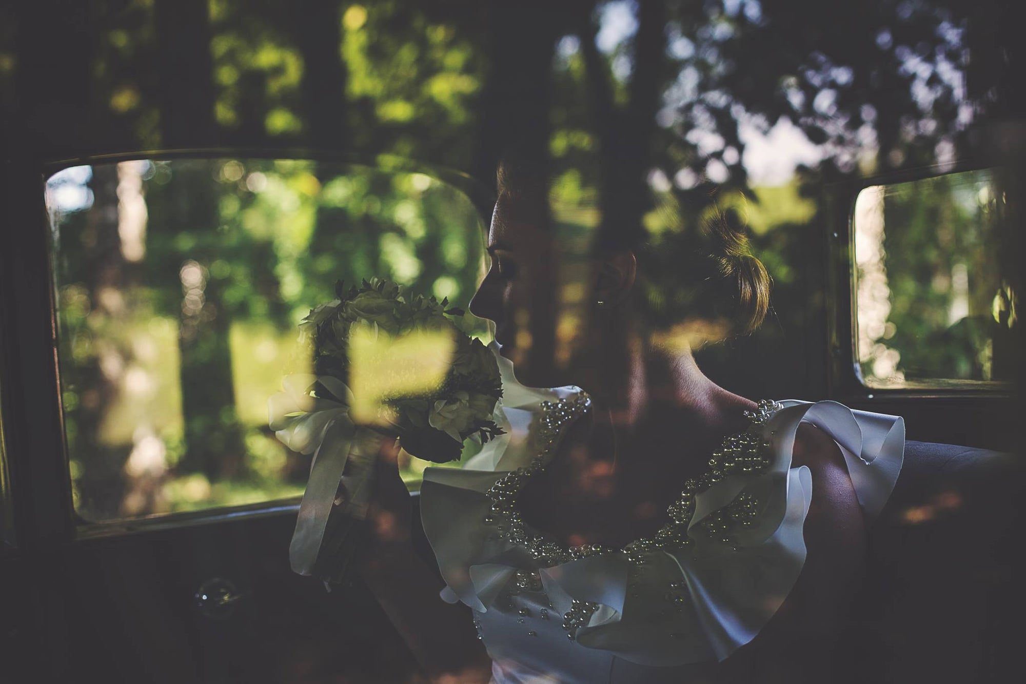 Bride in a pearl-embellished wedding dress by Krismil Bridal, holding her bouquet inside a vintage car, with soft reflections of greenery through the window.