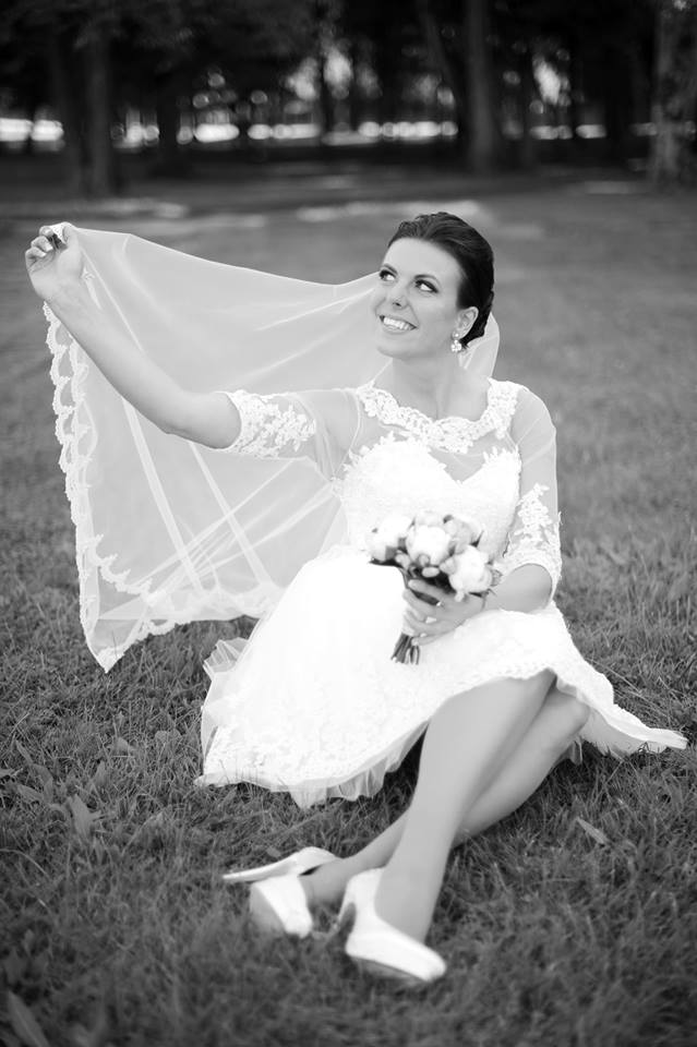 Bride in a short lace wedding dress by Krismil Bridal, sitting on the grass and holding her veil with a smile, while holding a bouquet in the other hand.