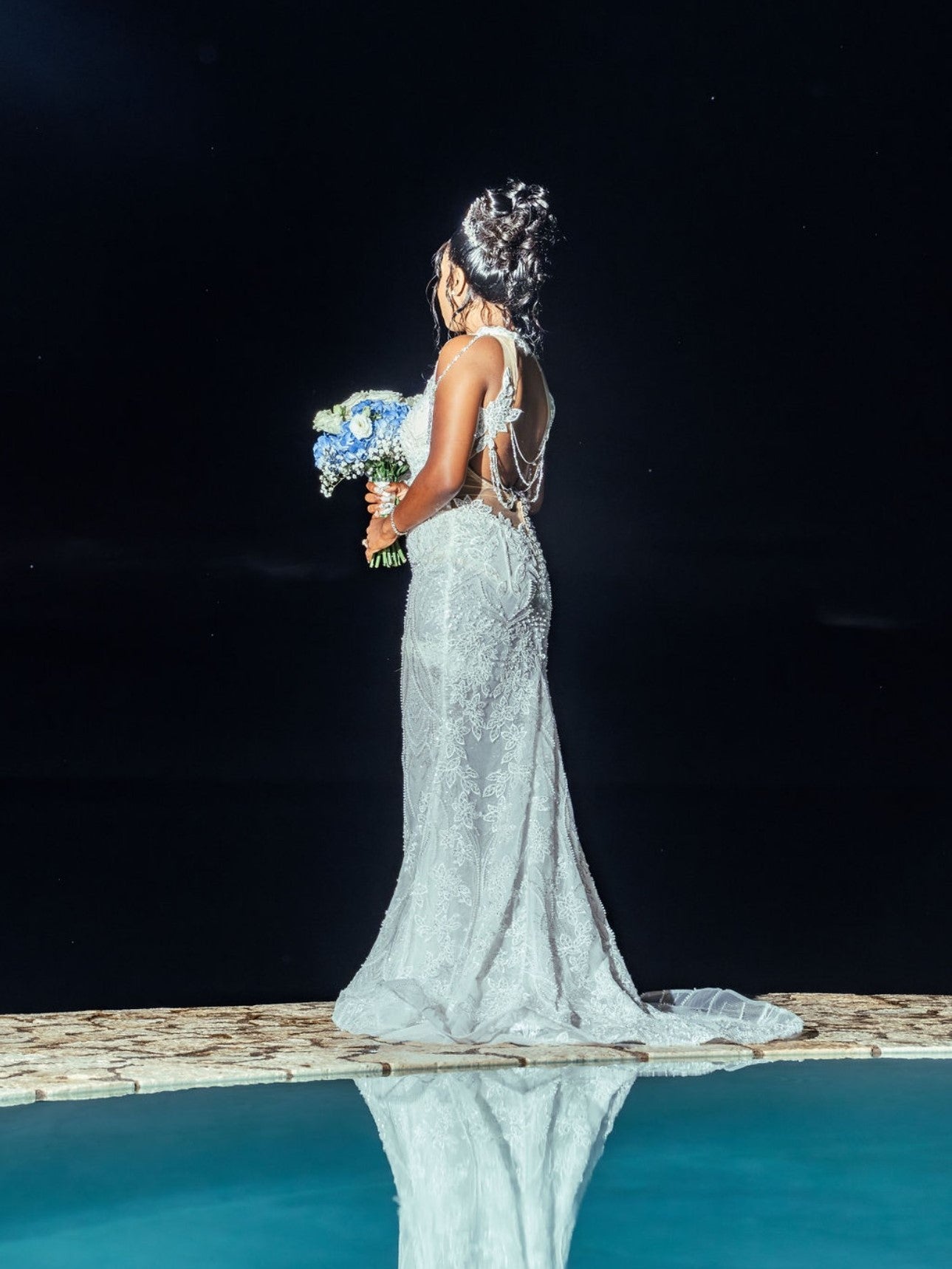 Bride Brittany in a beaded mermaid wedding dress by Krismil Bridal, showing the back of her gown while holding a blue and white bouquet at Ocean Cliff Hotel in Negril, Jamaica.