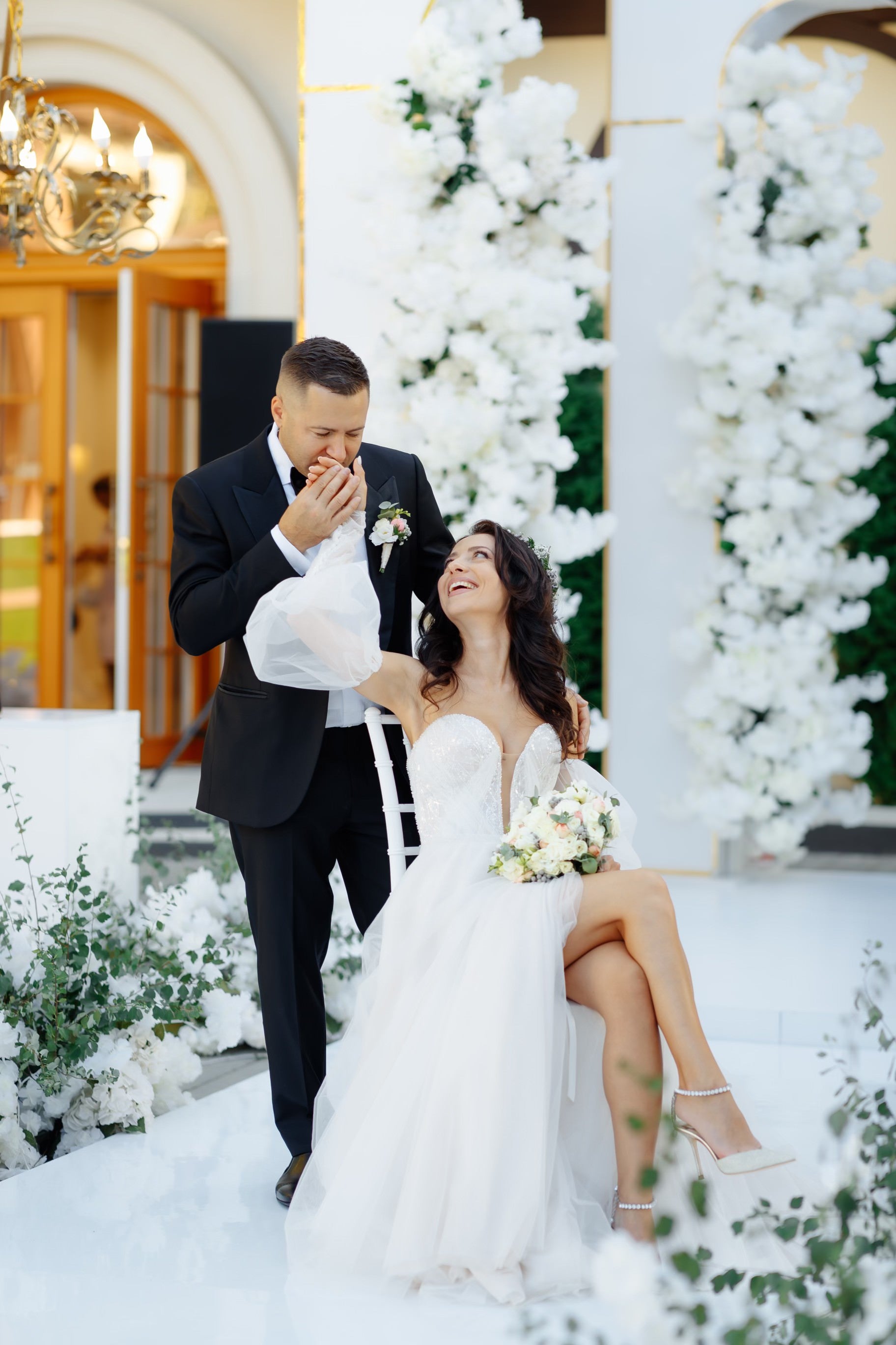 Groom Adrian kissing bride Doina's hand as she smiles, wearing an off-the-shoulder tulle wedding dress by Krismil Bridal.