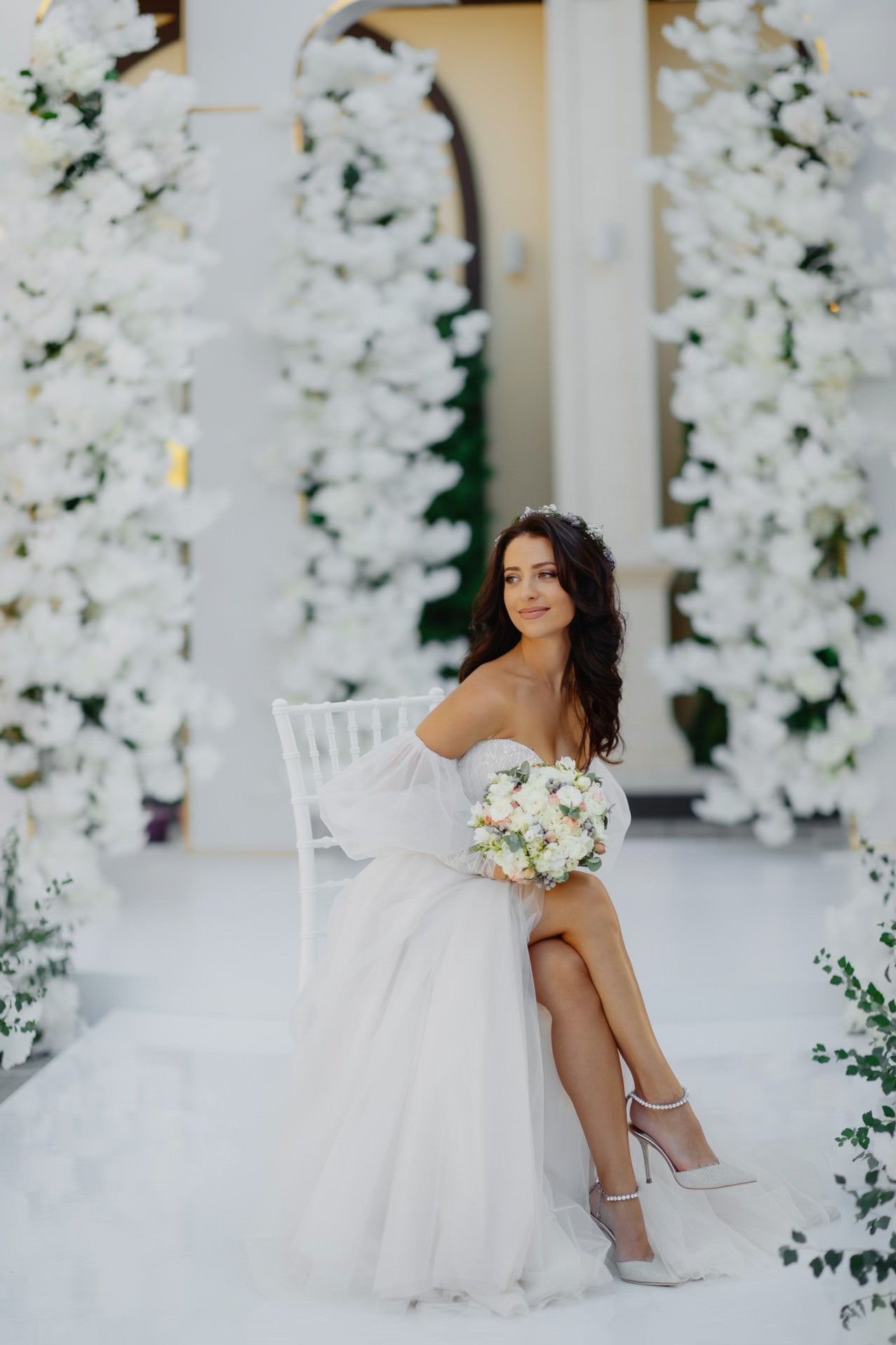 Bride Doina in an off-the-shoulder tulle wedding dress by Krismil Bridal, sitting with a bouquet and smiling.