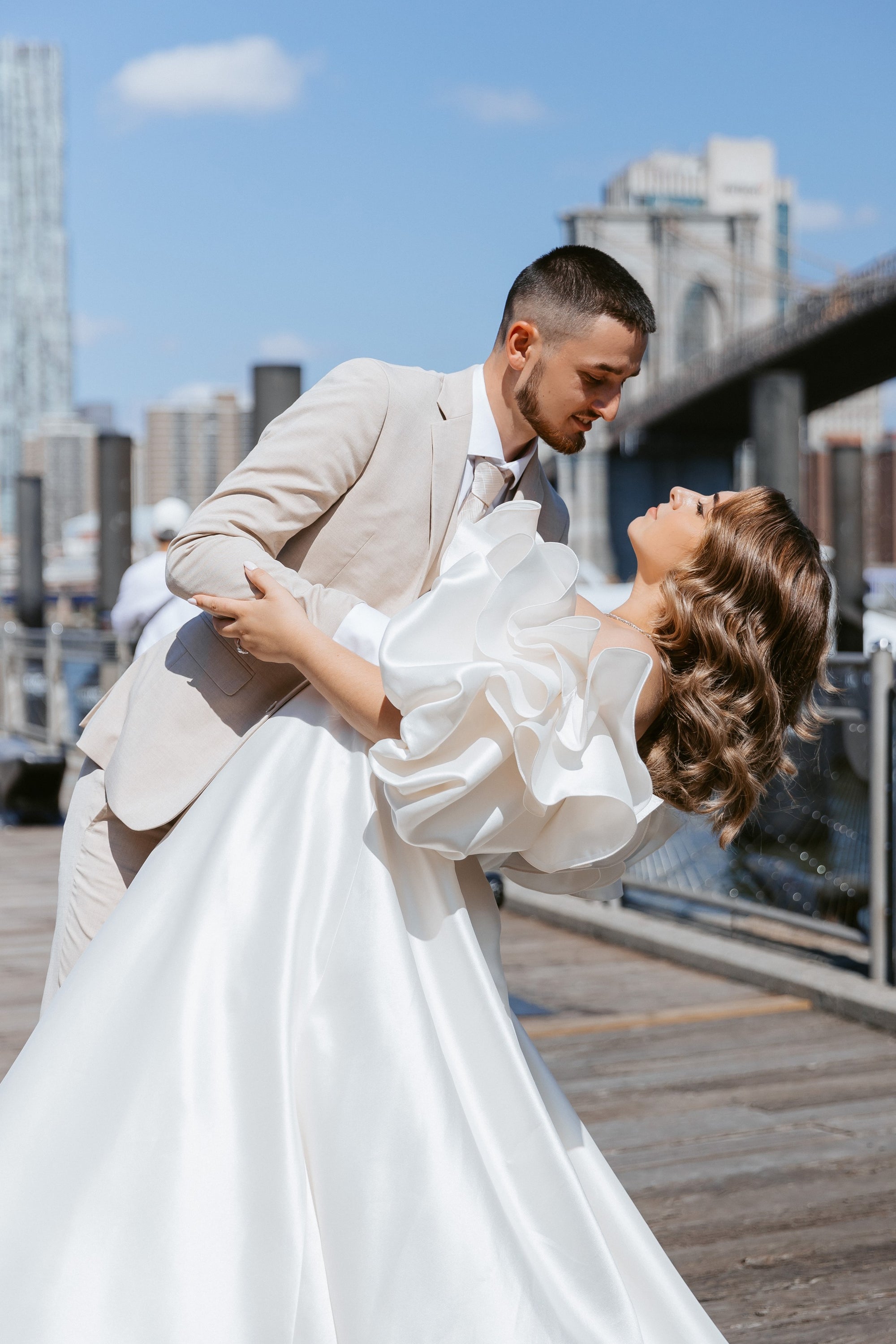 Bride Kristina in an off-the-shoulder A-Line Mikado wedding dress by Krismil Bridal, being dipped by her husband Andrii during their wedding photo shoot at Brooklyn Bridge Park.