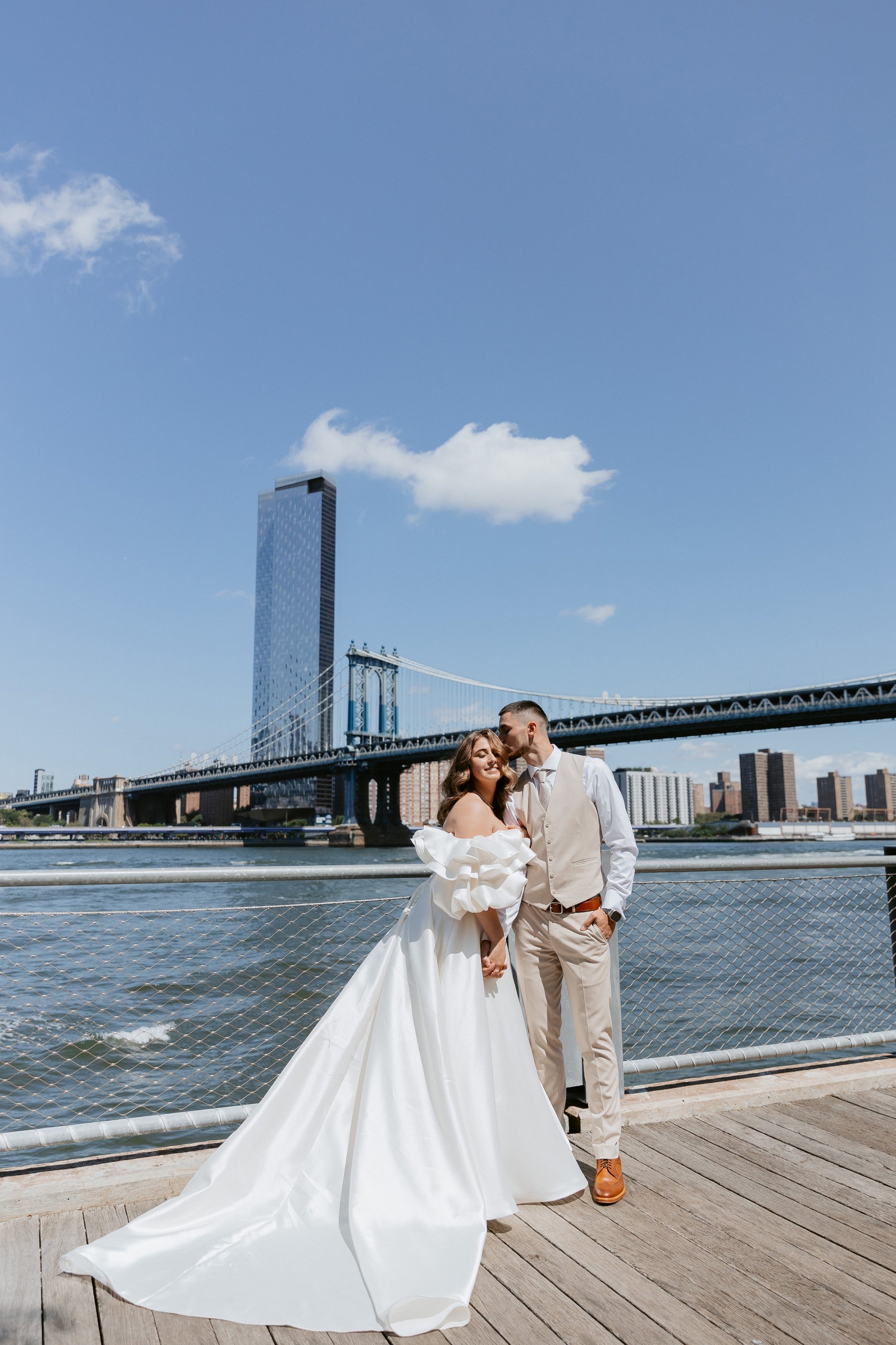 Bride Kristina in an off-the-shoulder Mikado gown by Krismil Bridal, posing with her husband Andrii at Brooklyn Bridge Park with the Manhattan Bridge in the background.