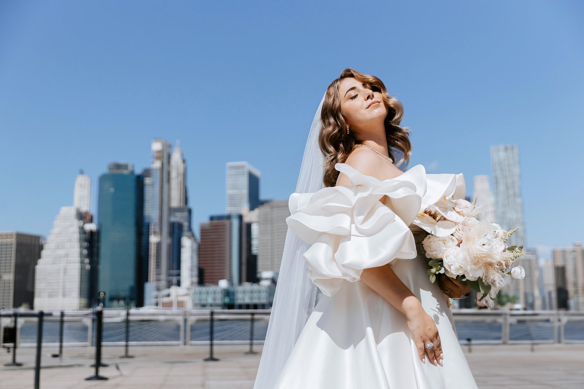 Bride Kristina in an off-the-shoulder A-Line Mikado wedding dress by Krismil Bridal, holding a bouquet with the New York City skyline in the background at Brooklyn Bridge Park.