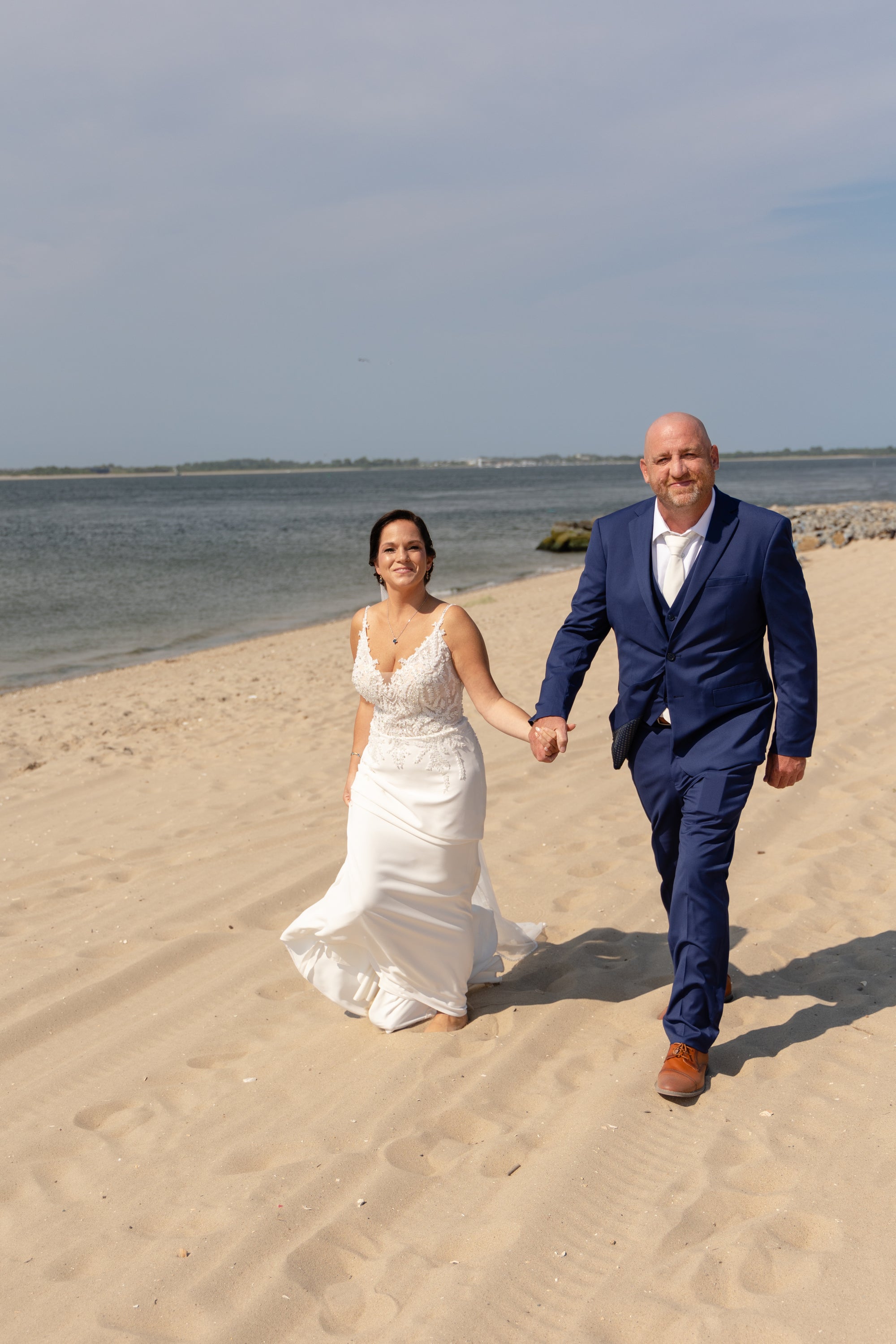 Bride Leigh Ann in a crepe mermaid wedding dress by Krismil Bridal, holding hands with her husband Kenneth while walking along the beach.