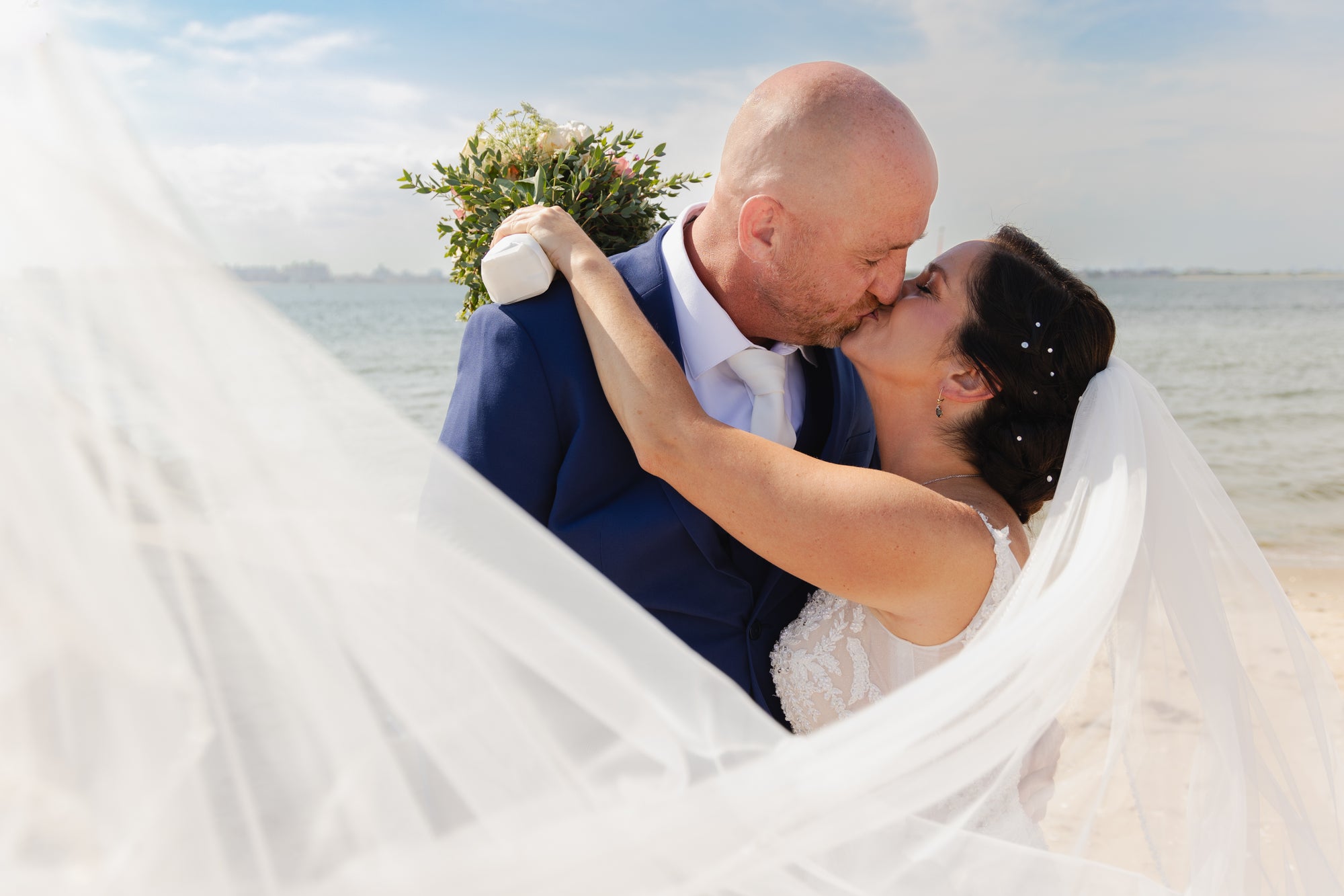 Bride Leigh Ann in a lace wedding dress by Krismil Bridal, kissing her husband Kenneth on the beach during their wedding.