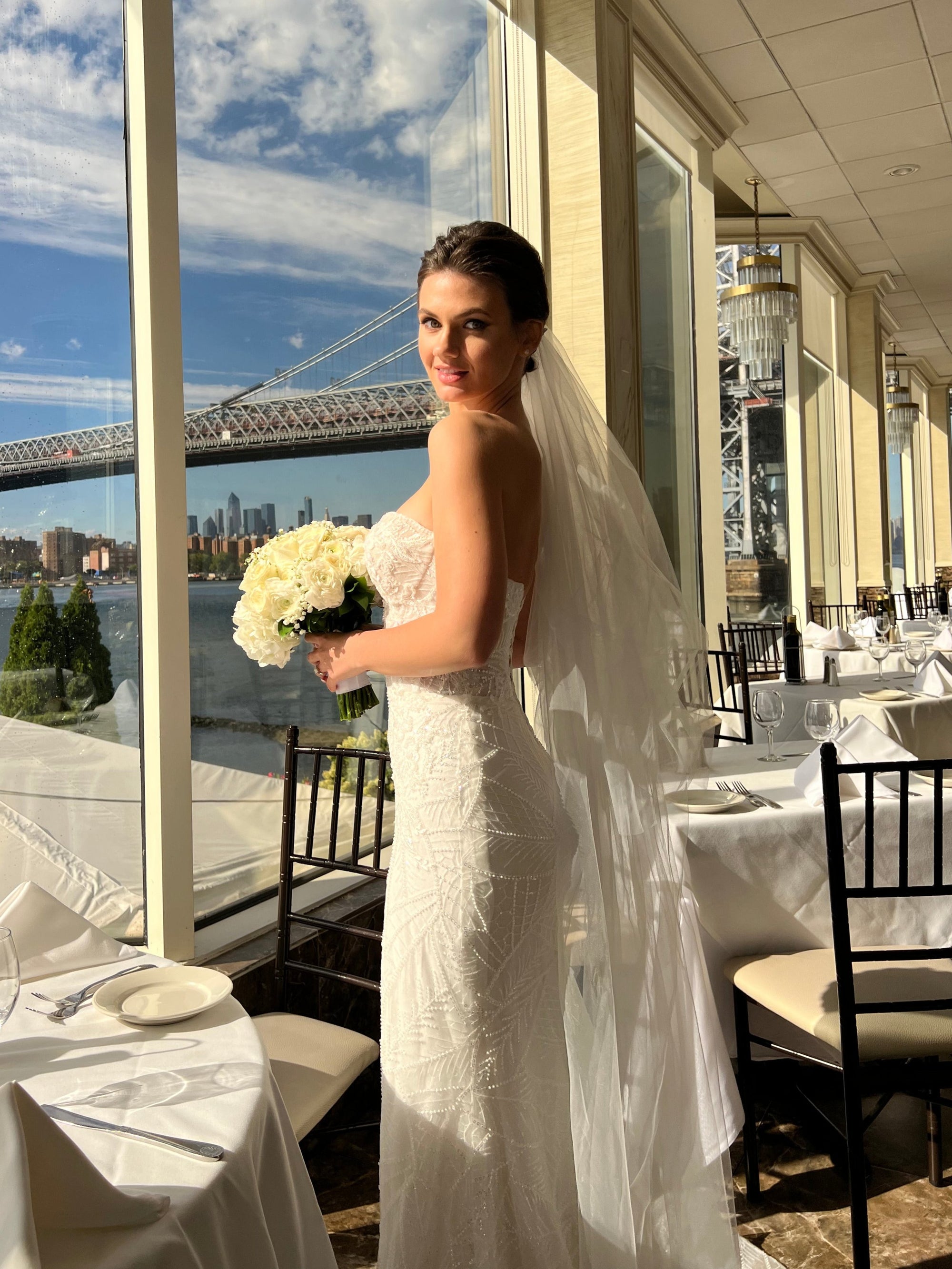 Bride Liliya in a beaded strapless wedding dress by Krismil Bridal, holding a bouquet of white roses and posing with a veil, with the river and bridge in the background.