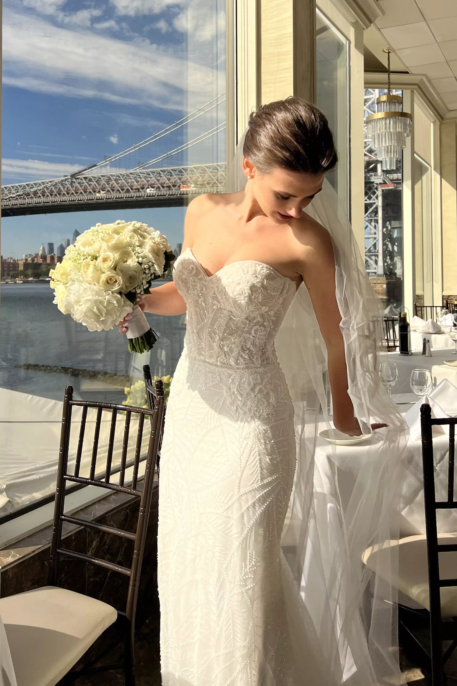 Bride Liliya in a strapless mermaid wedding dress by Krismil Bridal, holding a bouquet of white roses and posing indoors with a view of the bridge and river in the background.