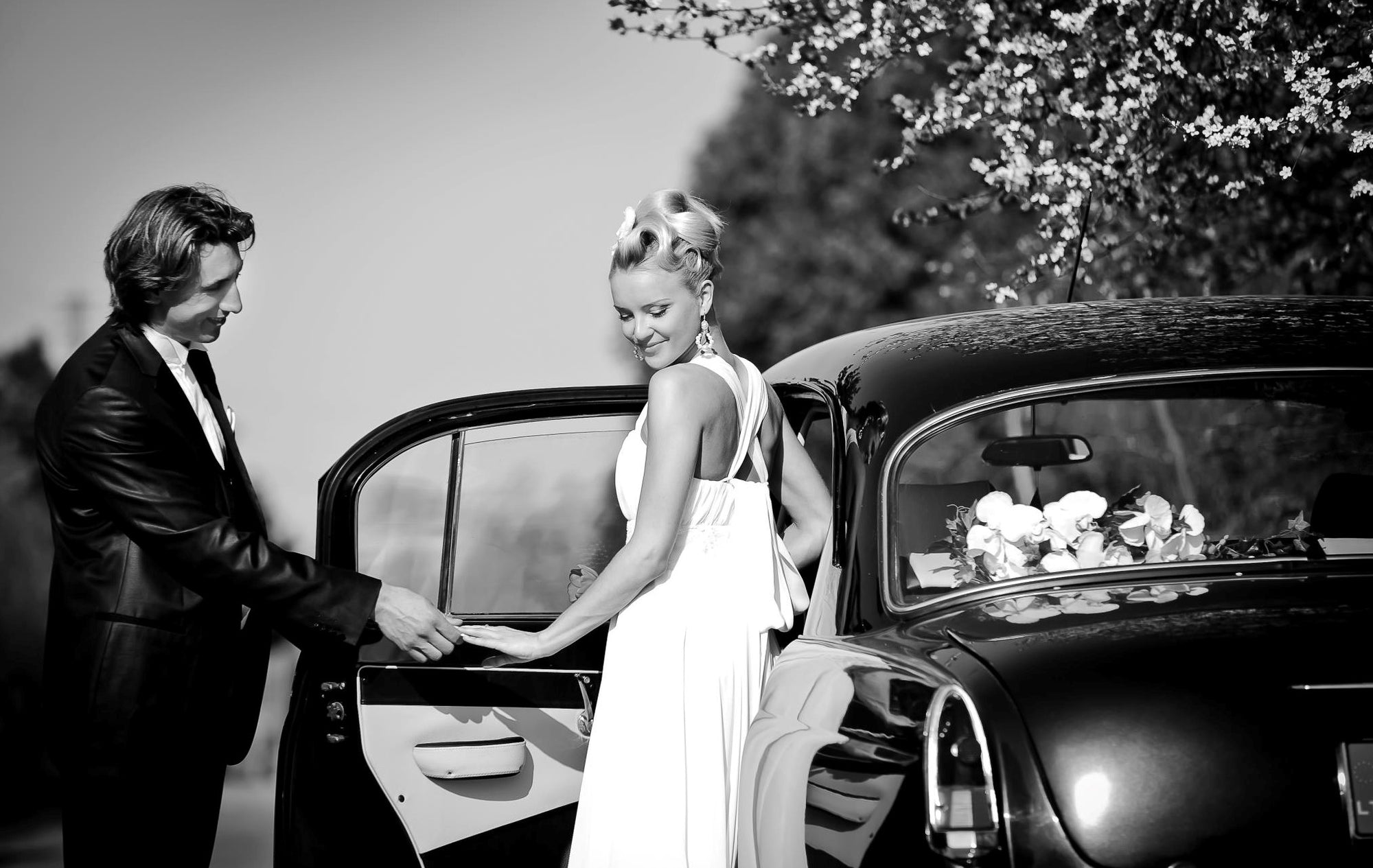 Bride stepping out of a vintage car wearing an elegant chiffon wedding dress by Krismil Bridal, helped by the groom during their wedding in Lithuania.