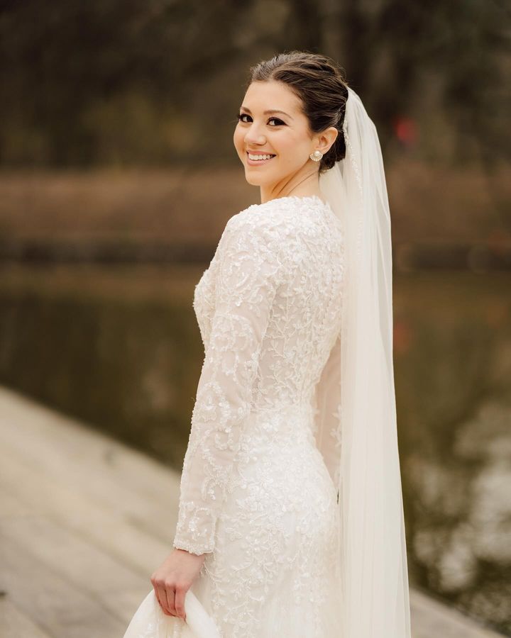 Bride Malka in a long-sleeve lace wedding dress by Krismil Bridal, smiling by a lakeside while wearing a veil.