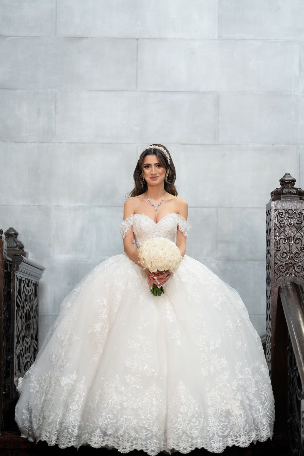 Bride Margaret in an off-the-shoulder lace ball gown wedding dress by Krismil Bridal, holding a bouquet while posing on a staircase in New York City.