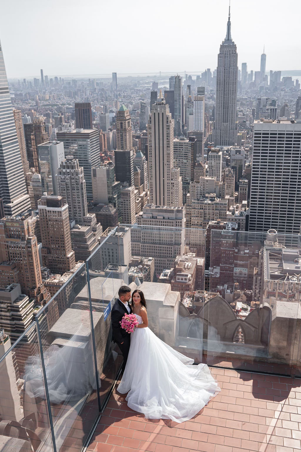 Bride Mariana in a strapless ball gown wedding dress by Krismil Bridal, holding a pink bouquet with her groom at Top of the Rock Observation Deck in New York City.
