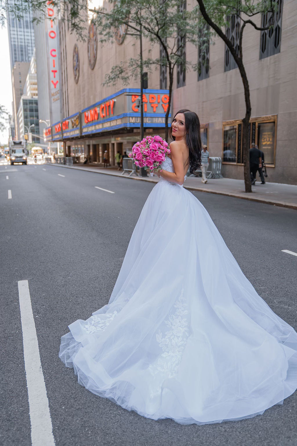 Bride Mariana in a tulle ball gown wedding dress by Krismil Bridal, holding a pink bouquet in front of Radio City Music Hall in New York City.
