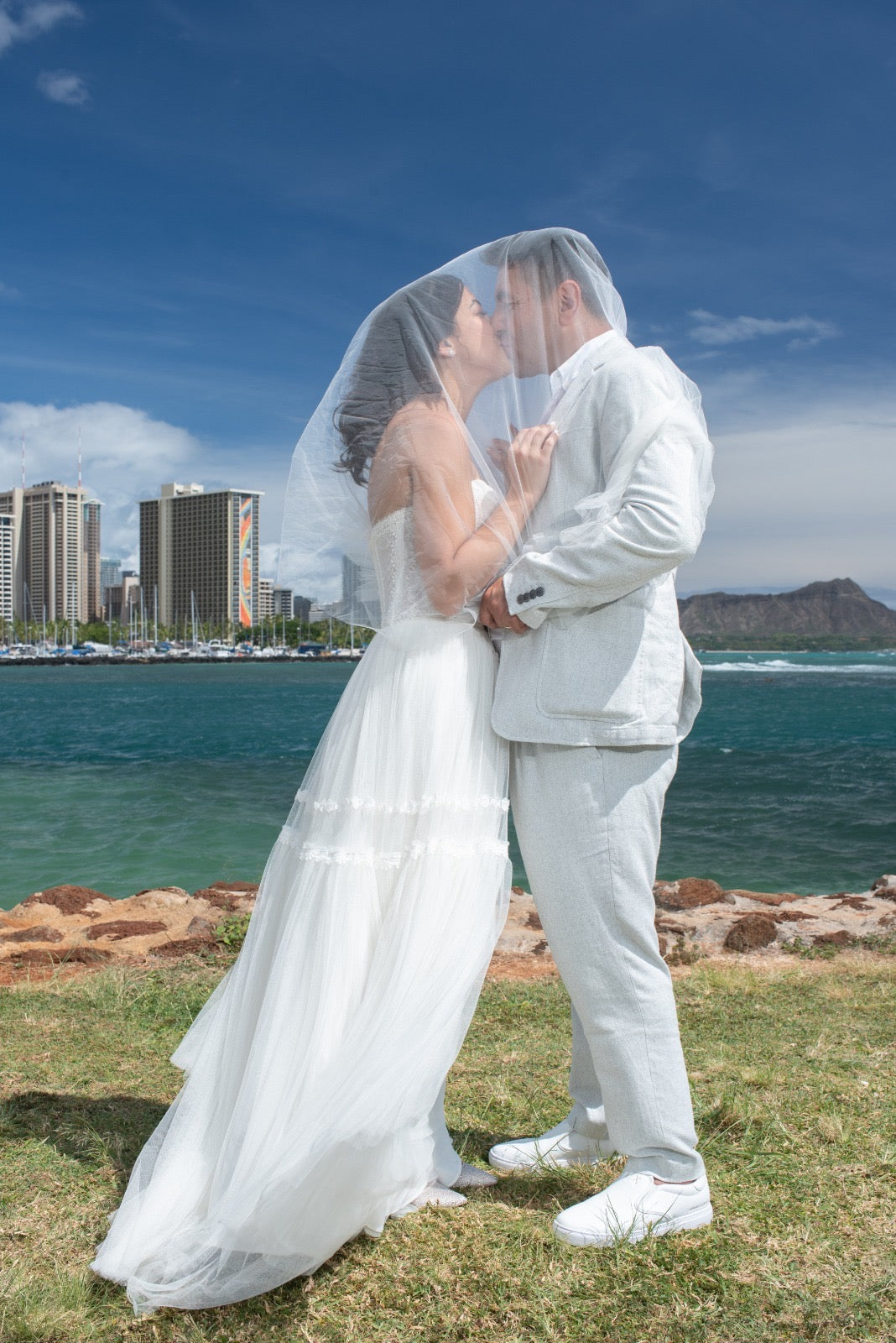 Bride Melis in a boho wedding dress by Krismil Bridal sharing a kiss with the groom under her veil, with the ocean and city skyline in the background during their wedding in Hawaii.