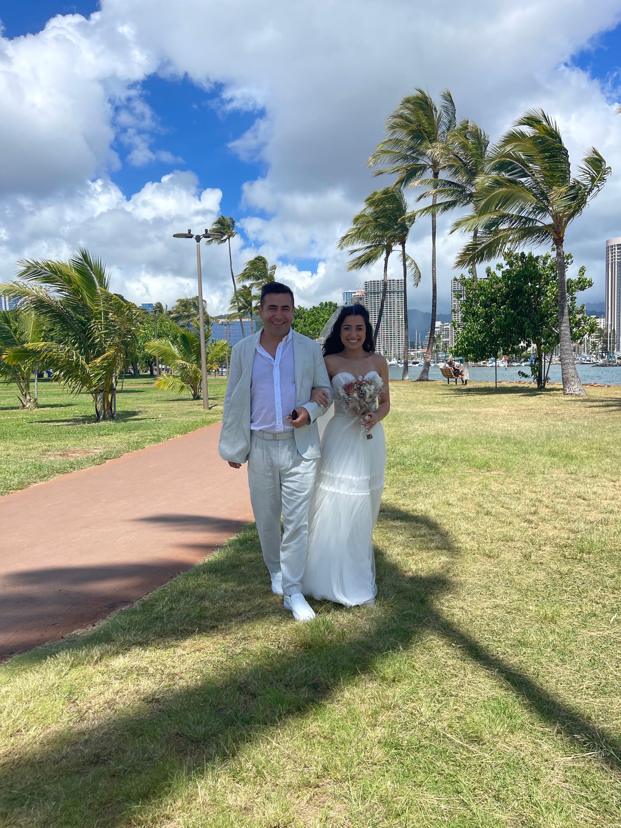 Bride Melis in a strapless A-line wedding dress by Krismil Bridal walking with the groom along a palm-lined path during their sunny outdoor wedding in Hawaii.