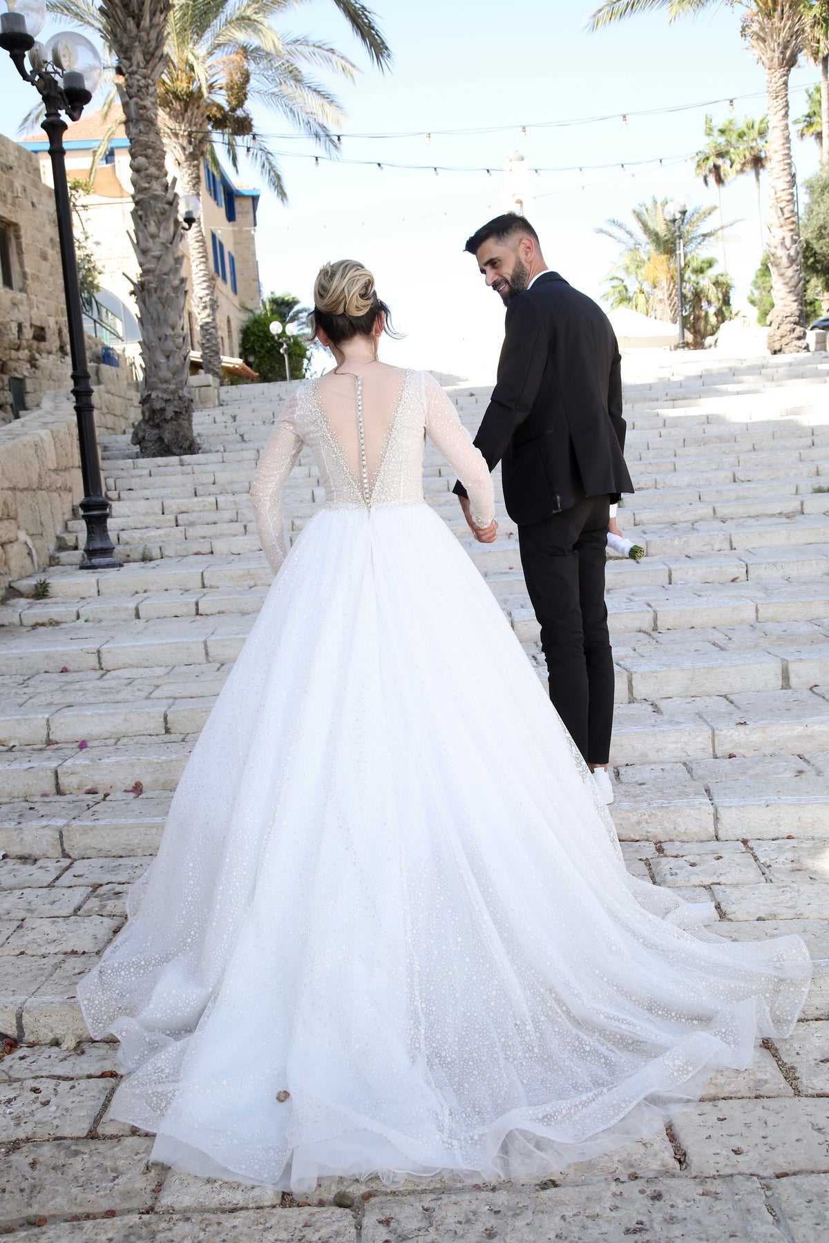 Bride Melissa in a sparkly ball gown wedding dress by Krismil Bridal walks hand in hand with Groom David up stone stairs in Israel, as he lovingly looks back at her.
