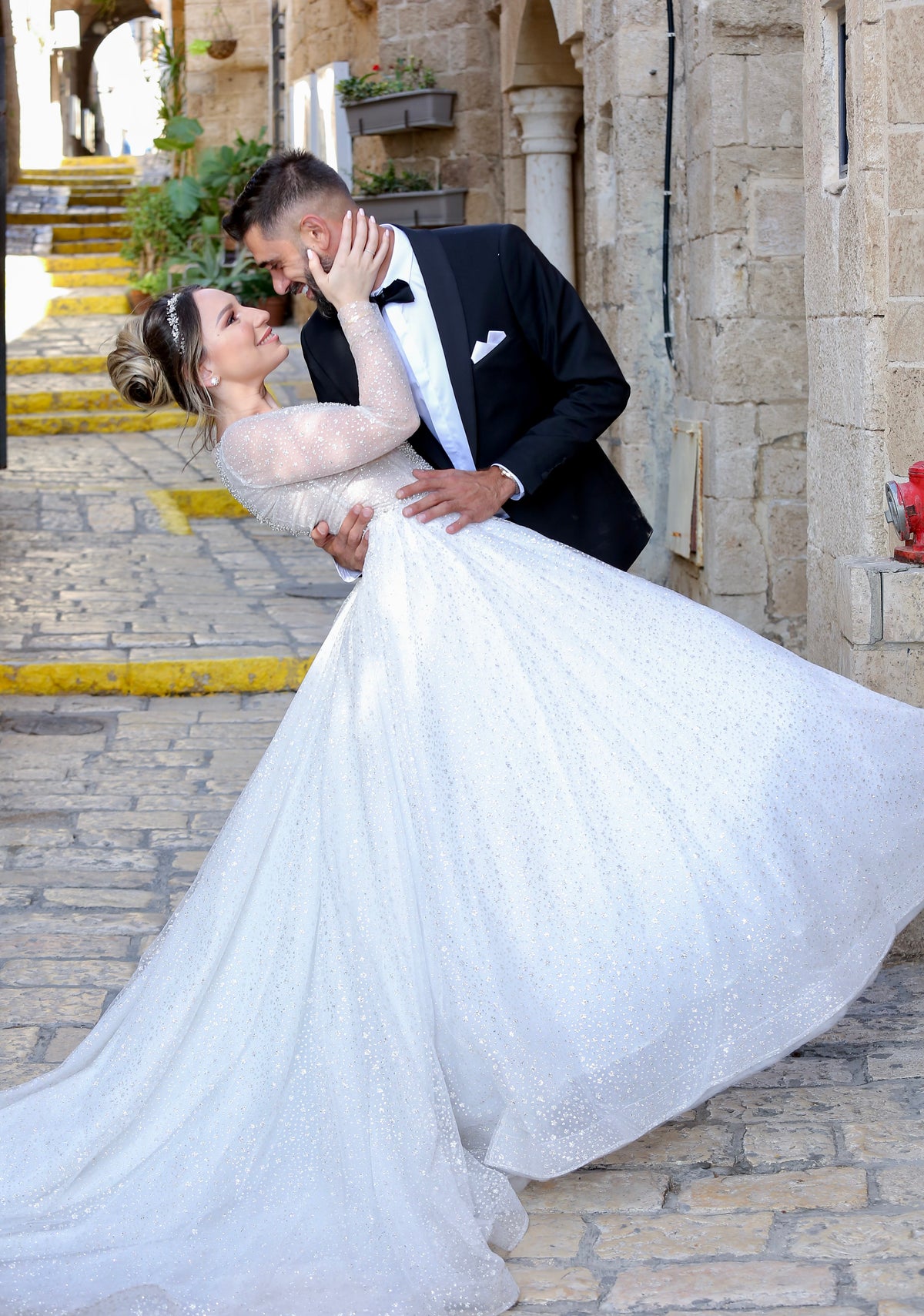 Bride Melissa in a sparkly long-sleeved gown by Krismil Bridal shares a romantic dance moment with Groom David on the cobblestone streets of Israel, both smiling and glowing with love.