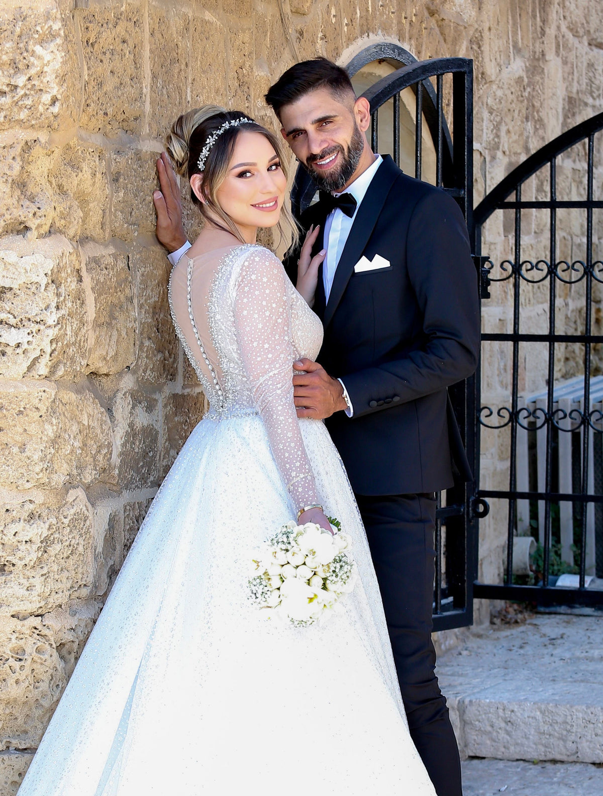 Bride Melissa in a sparkly long-sleeved wedding dress by Krismil Bridal, standing with Groom David against a stone wall during their wedding in Israel, both smiling and radiating happiness.