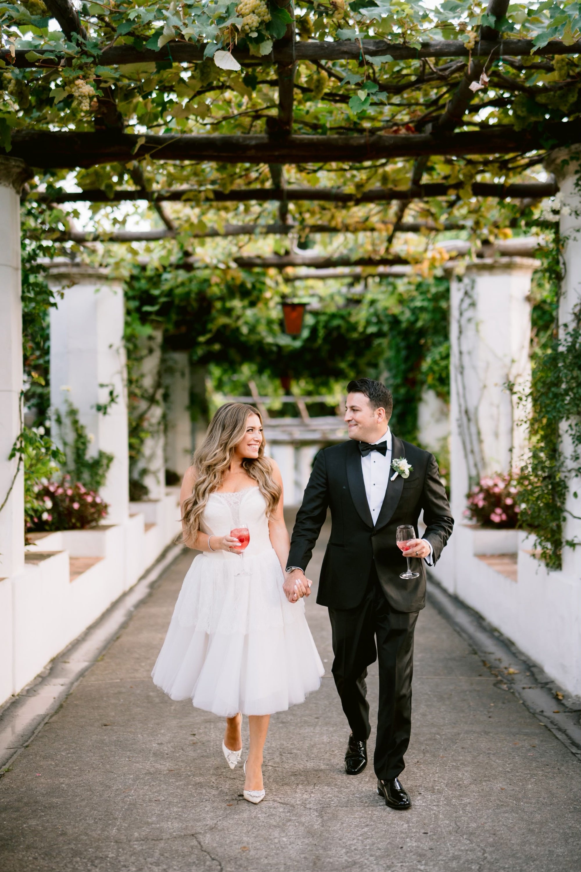 Bride Michelle and groom walking hand in hand with drinks during their wedding in Ravello, Italy, surrounded by a lush vine-covered walkway.