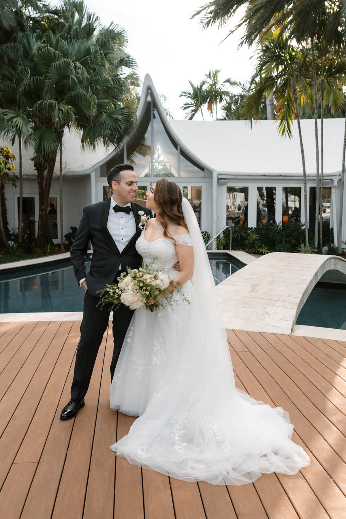 Bride Penina in an off-the-shoulder lace wedding dress by Krismil Bridal, standing poolside with her husband David at their wedding in Miami, Florida.