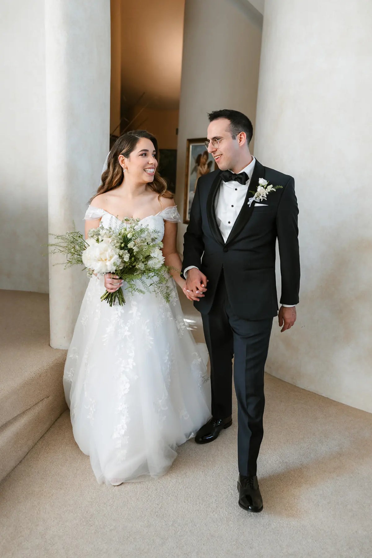 Bride Penina in an off-the-shoulder lace wedding dress by Krismil Bridal, walking hand-in-hand with her husband David at Hibiscus Island in Miami.