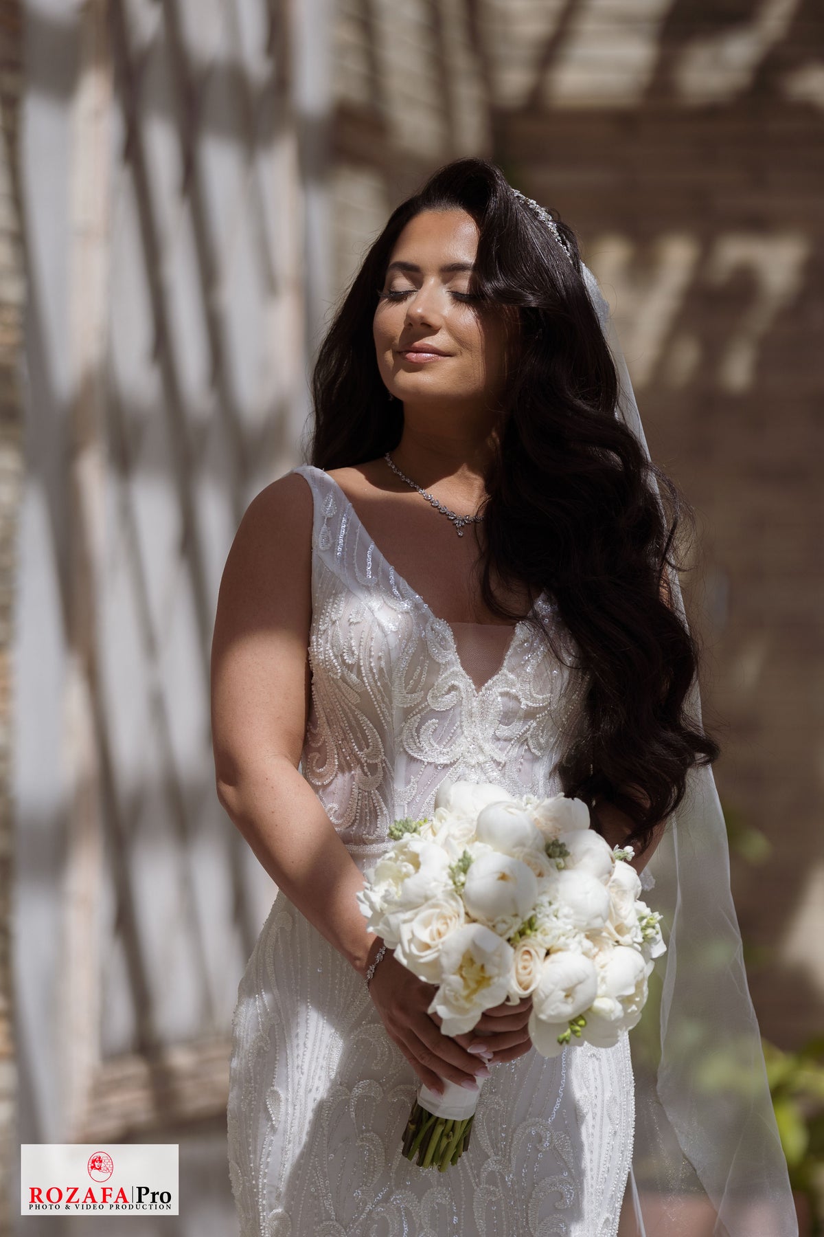 Bride Rochelle in a beaded mermaid wedding dress by Krismil Bridal, holding a bouquet and standing in the sunlight at Untermyer Gardens Conservancy.