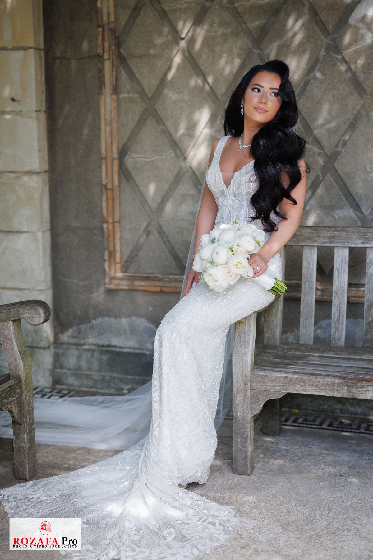 Bride Rochelle in a beaded mermaid wedding dress by Krismil Bridal, sitting on a bench and holding a bouquet at Untermyer Gardens Conservancy.