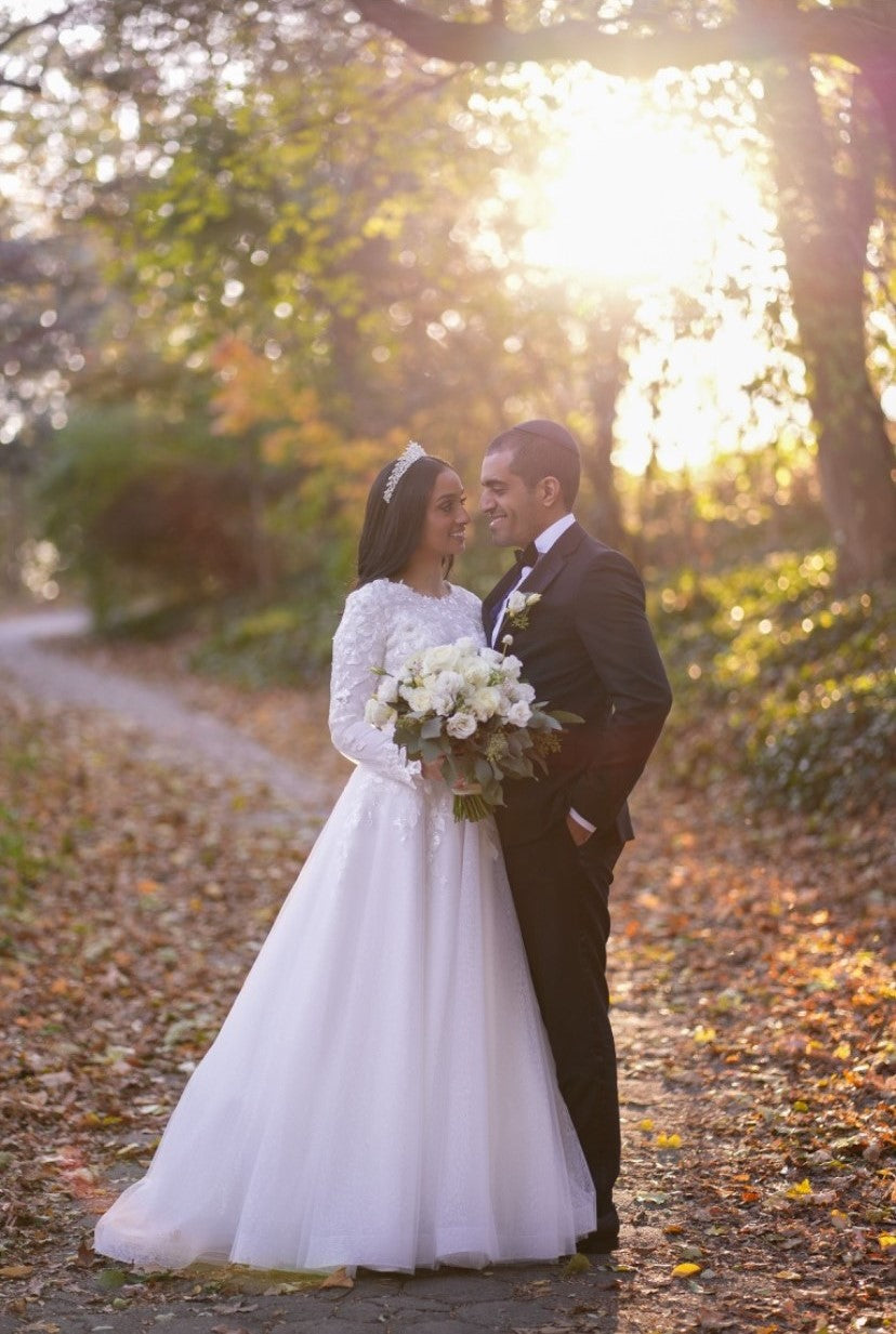 Bride Tohar wearing a modest, long-sleeved wedding dress by Krismil Bridal, holding a bouquet and standing with her groom in a serene forest setting during their religious wedding.