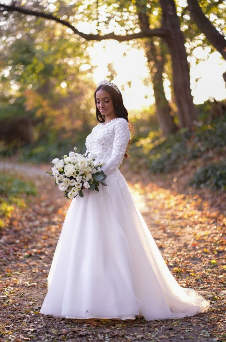 Bride Tohar in a modest lace A-line wedding dress by Krismil Bridal, holding a bouquet of white flowers and standing in a sunlit forest path, with soft golden light illuminating her on her special day.