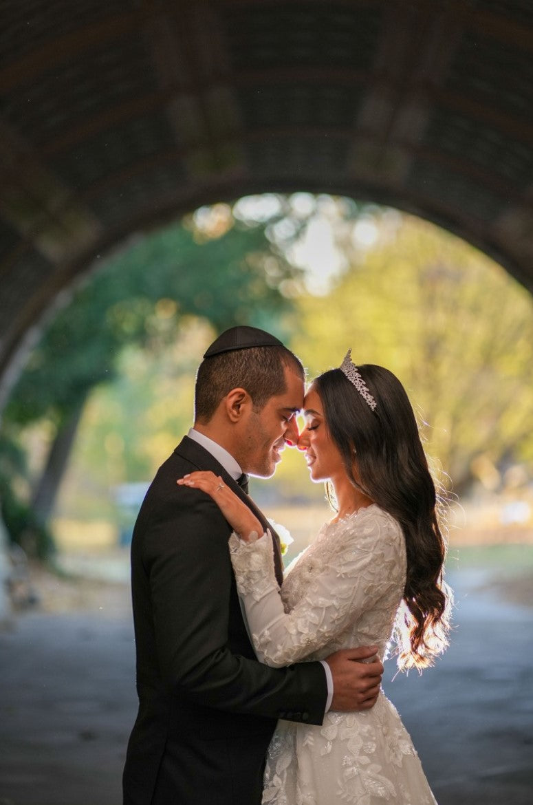 Bride Tohar in a modest, long-sleeved wedding dress by Krismil Bridal, embracing her groom under a beautifully lit tunnel, with the soft glow of sunlight illuminating their faces during their intimate wedding moment.