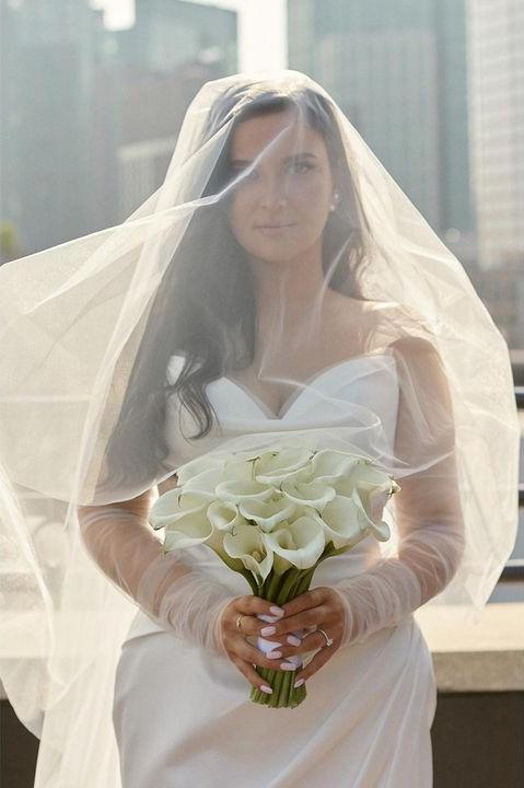 Bride Valerie in a satin mermaid wedding dress by Krismil Bridal, holding a calla lily bouquet and wearing a veil over her face.