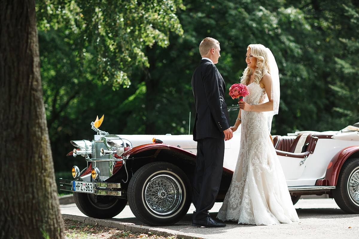Bride in a lace mermaid wedding dress by Krismil Bridal and groom standing next to a vintage car during their wedding in Lithuania.